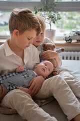 Obraz premium Three brothers sitting together on a bed at home, older siblings gently holding a newborn baby, soft natural light and calm interior