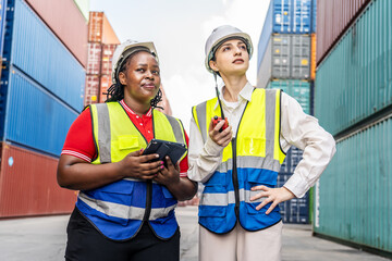 Two diverse female logistics workers in safety vests and hard hats using digital tablet and radio at shipping port terminal. Professional team discussing cargo operations in container yard background