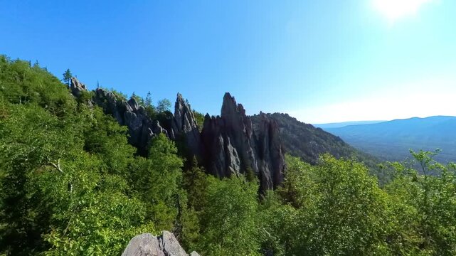 summer landscape shot from a high vantage point, capturing the dramatic, sharp peaks of rock formations rising above a vibrant green forest under a clear blue sky in Taganay National Park, near Zlatou