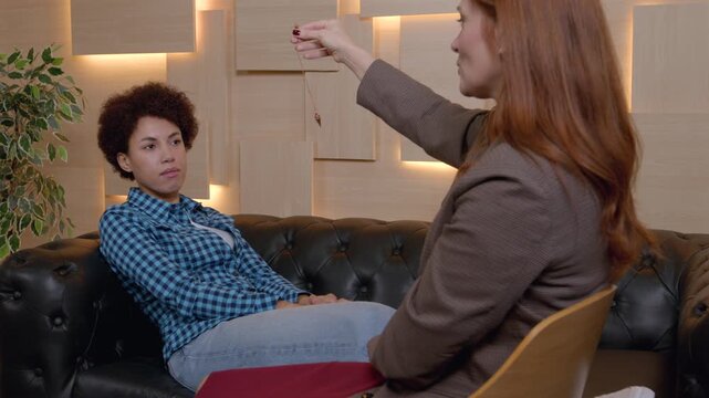 Mature woman psychologist hypnotizing pretty African American female patient using pendulum, to create state of focused attention and increased suggestibility in treatment of psychological disorder.