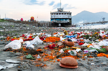 Pollution and religious waste litter a riverbank in India, with discarded offerings, plastic debris, and flowers scattered along the shore near a docked vessel