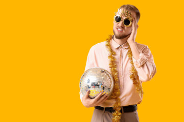 Young man with hangover and disco ball after Birthday party on yellow background
