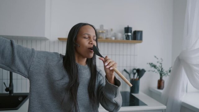 Pretty young woman enjoying music and singing son at home kitchen, good mood. Positive black lady having fun alone at home in weekend, be happy and feel joy, funny female person with kitchenware