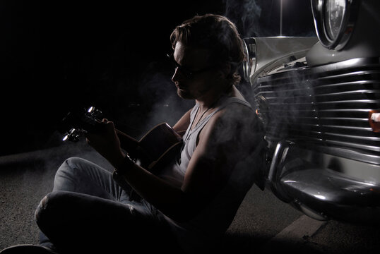 Man playing guitar at night next to oldtimer car with smoke outdoors