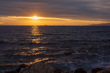 Moody sunset scene with golden light reflecting on dark Aegean Sea waves under layered clouds. Peaceful seascape near Athens, Greece, suitable for travel, nature and background use.