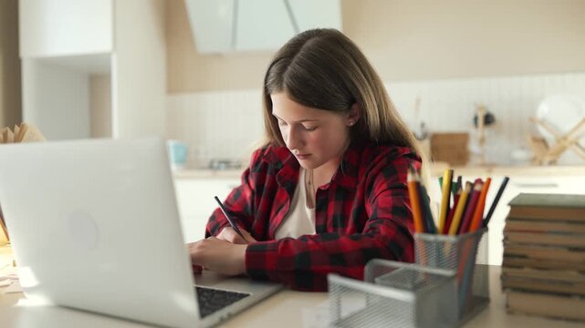 Girl studying at desk writing in notebook and using laptop while pencil moves across paper student teenager doing homework at home with book stack pencil cup lamp study space concentration