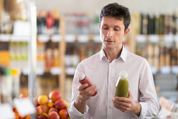 Man in light shirt took some varieties of fruit smoothie from shelf. Client compare fruit and berry fresh mix items package. Customer examines contents of package, choose smoothie © JackF