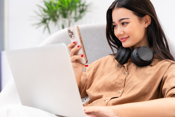 Young beautiful, stylish, happy Asian woman with headphone on neck holding, looking at smartphone, smiling, with computer laptop, video calling. Digital technology, modern lifestyle, online learning