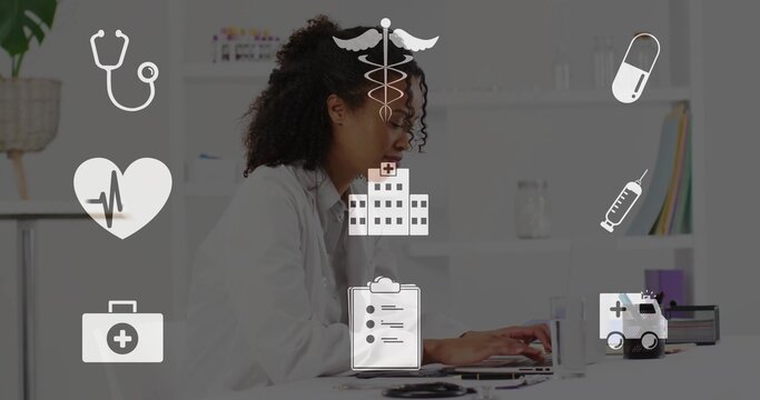 Working woman in white lab coat typing on laptop at clinical office desk, with stethoscope nearby