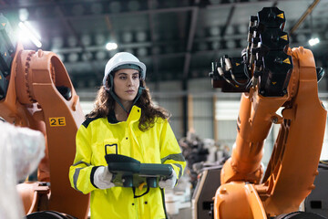 Female engineer wearing safety helmet and high visibility jacket operates robotic arm using digital tablet in advanced manufacturing facility. Concept of automation and smart industry.