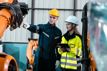 Male and female engineers in safety uniforms collaborate using digital tablets to control robotic arm equipment in smart factory. Concept of teamwork, automation and industry innovation.