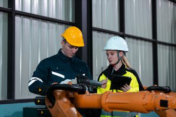 Male and female engineers in safety uniforms collaborate using digital tablets to control robotic arm equipment in smart factory. Concept of teamwork, automation and industry innovation.