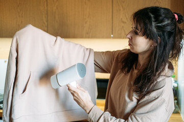 Woman uses portable garment steamer on sweatshirt in kitchen. Lightweight travel gear, business trip essentials, garment care.