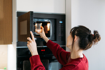 Woman reaches into microwave to heat glass of milk in modern kitchen. Heating oat milk, vegan latte preparation, dairy free lifestyle.