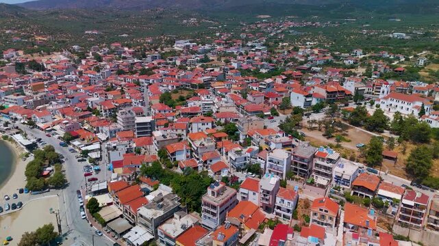 Aerial drone flight over the harbor and coastal town of Limenaria on Thasos Island, Greece, highlighting the rooftops and marina under a bright summer sky.