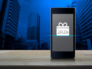 Gift box happy new year 2026 icon on modern smart mobile phone screen on wooden table over office city tower and skyscraper, Business happy new year 2026 shop online concept