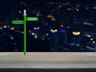 2026 and 2025 direction sign plate with green pencil on wooden table over blur colorful night light of modern office city tower and skyscraper, Business happy new year 2026 planning concept