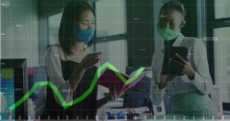 Green graph moving over two diverse female office colleagues wearing face masks discussing at office - Powered by Adobe