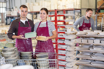 Smiling successful male and female artisans wearing maroon aprons, proudly displaying crafted ceramic plates in spacious workshop