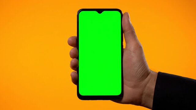 Hand holding smartphone with green screen display against vibrant orange background for mockup