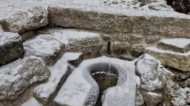 stone fountain, a spring in the winter snow-covered from large stone blocks. Water flows out of a small pipe and flows into a central, horseshoe-shaped stone bowl. covered with a thin layer of snow, 