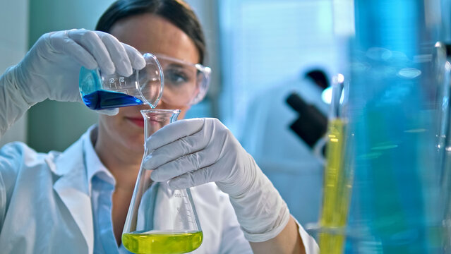 A scientist in a lab coat and gloves carefully pours a blue liquid from a beaker into a flask containing a yellow substance, in a well-lit lab with various chemical containers.
