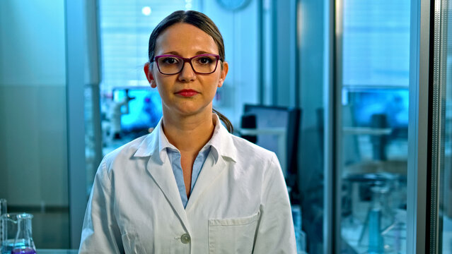A focused female scientist in a lab coat and glasses stands in a modern lab with computer monitors and scientific equipment, suggesting a setting dedicated to medical research or chemical analysis.