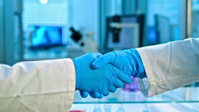 Two scientists in lab coats and blue gloves shake hands in a lab. Background includes scientific equipment like monitors and glassware, indicating collaboration in medical research.