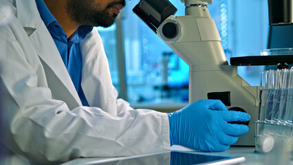 A scientist in a lab coat and blue gloves examines a sample under a microscope in a well-lit lab with test tubes and scientific equipment.