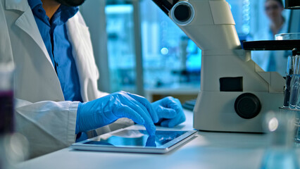 A scientist in a lab coat and blue gloves uses a tablet to analyze data beside a microscope in a modern lab, emphasizing medical research and healthcare innovation. The scene highlights meticulous.