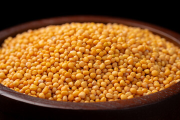 Pile of yellow mustard seeds in a brown wooden bowl on a black background.