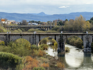 Fototapeta premium Serpis River with high water flow in Gandia, Spain. Includes stone arch bridge, lush green vegetation, and distant mountains