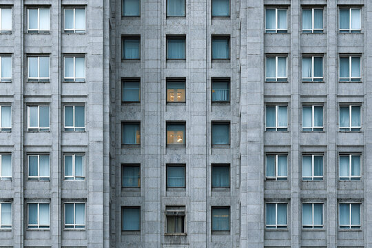 Corporate skyscraper facade texture with evenly spaced rectangular windows in gray stone, some windows lit warmly inside, creating calm urban architectural pattern