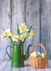 Easter holiday greeting card template; bouquet of white daffodils in green vintage coffee pot and wicker basket with easter eggs on a wooden background; copy space