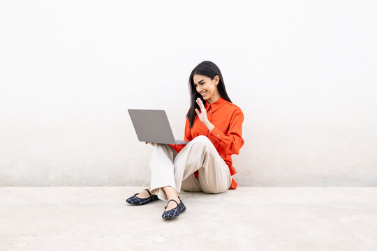 Confident woman in orange shirt sitting on the floor with laptop, smiling and saying hello