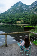 A woman sits in a chair overlooking a lake. The water is calm and the sky is cloudy. The woman is enjoying the view and the peaceful atmosphere