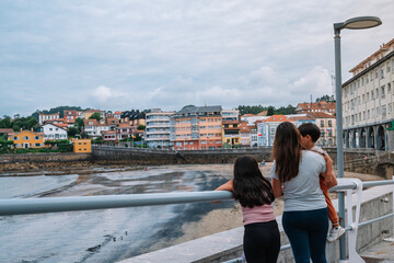 A woman and two children stand on a pier overlooking a body of water in Luanco, Asturias. The sky is cloudy and the atmosphere is calm and peaceful.