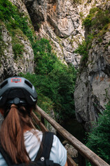 A woman wearing a helmet is looking down a narrow path. The path is surrounded by trees and rocks