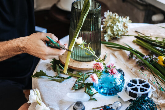 Hands use green snips to trim thick plant stem on table next to green glass vase. Zero-waste floristry, sustainable floral foam alternatives, eco-friendly flower arranging.