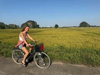 Naklejka premium Young woman smiling at camera cycling on rural road beside golden rice field paddies and karst peaks Vietnam