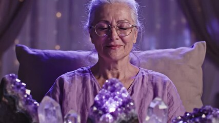 Senior woman meditating with large amethyst crystals and quartz. Elderly spiritual healer sitting in a mystical purple room with bokeh lights. Esoteric magic and crystal healing concept