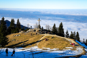 The summit cross on the Kampenwand, a striking mountain in the Chiemgau Alps. The mountain landscape lies above a thick cloud cover under a bright blue winter sky.