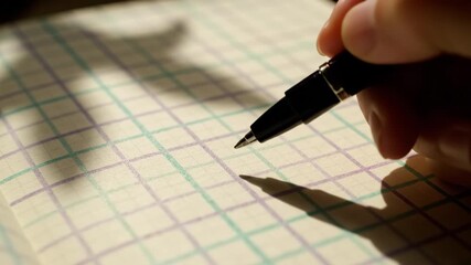 Handheld pen over grid paper with warm light and soft shadow, close up of hand writing checkered notebook page showing tactile paper texture, graph paper grid shadow across notebook with shallow