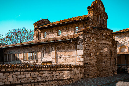 Sun-drenched Byzantine church with terracotta roofs and layered masonry under a cyan sky. Its ancient stone arches and rustic walls glow warmly on a quiet, historic cobblestone street.