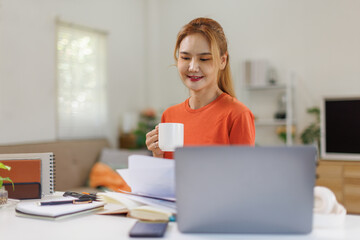 Asian businesswoman at home office reads task list on laptop drinks coffee takes sip smiles stays focused. Freelancer girl at table enjoys cup gets energy plans steps types reply and saves file.
