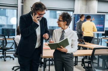 Business professionals engaged in conversation while using smartphone and notebook in modern office environment, showcasing collaboration and teamwork in a corporate setting