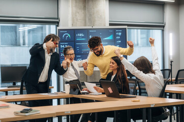 Group of diverse professionals celebrating success in a modern office, with digital screens displaying data and graphs, showcasing teamwork and achievement