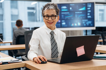 Businesswoman with short gray hair is smiling while working on laptop in modern office, showcasing a collaborative workspace with technology and data displays