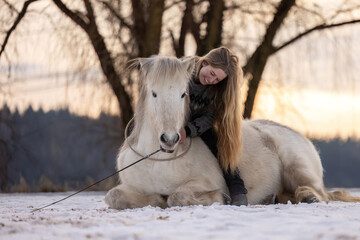 Emotional moment of trust between a woman and an Icelandic horse lying together on a snowy field at sunset
