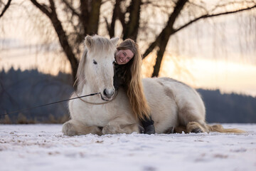 Emotional moment of trust between a woman and an Icelandic horse lying together on a snowy field at sunset